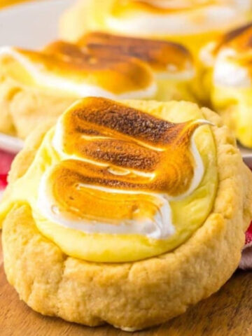 A wooden board features cookies topped with toasted meringue. A plate in the background holds more cookies. A red and white cloth is partially visible underneath.