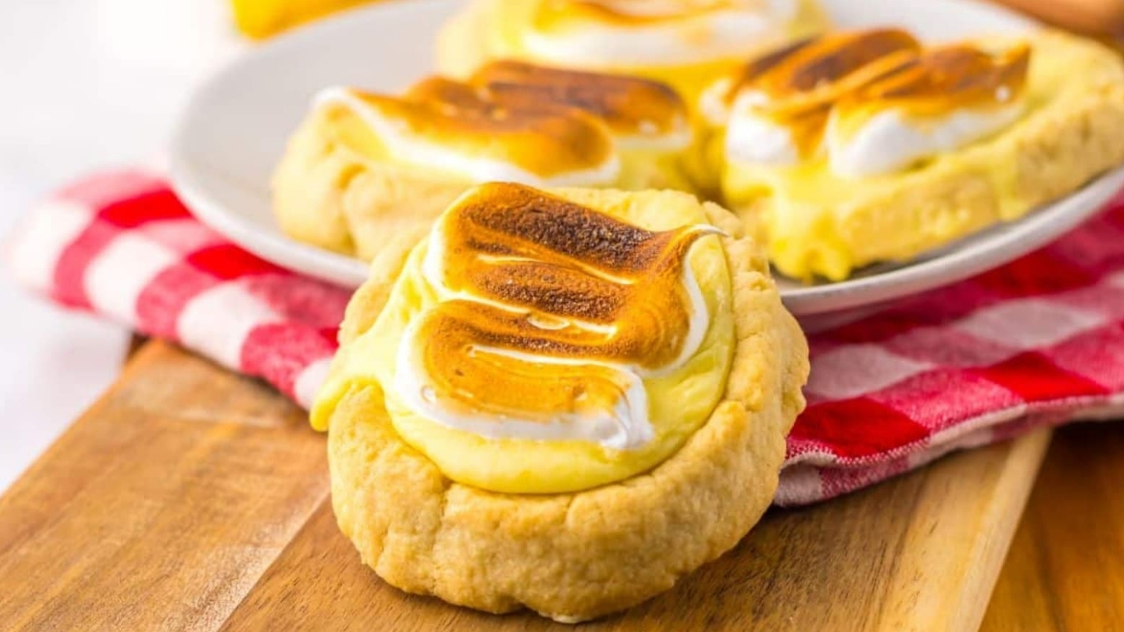 A wooden board features cookies topped with toasted meringue. A plate in the background holds more cookies. A red and white cloth is partially visible underneath.