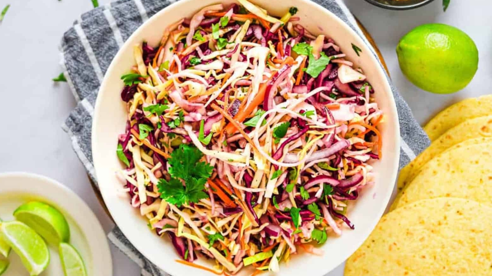 Bowl of colorful coleslaw with shredded cabbage, carrots, and cilantro, next to lime wedges and tortillas.