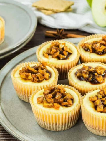 Mini cheesecakes topped with caramelized apples on plates, with a green apple and graham crackers in the background.