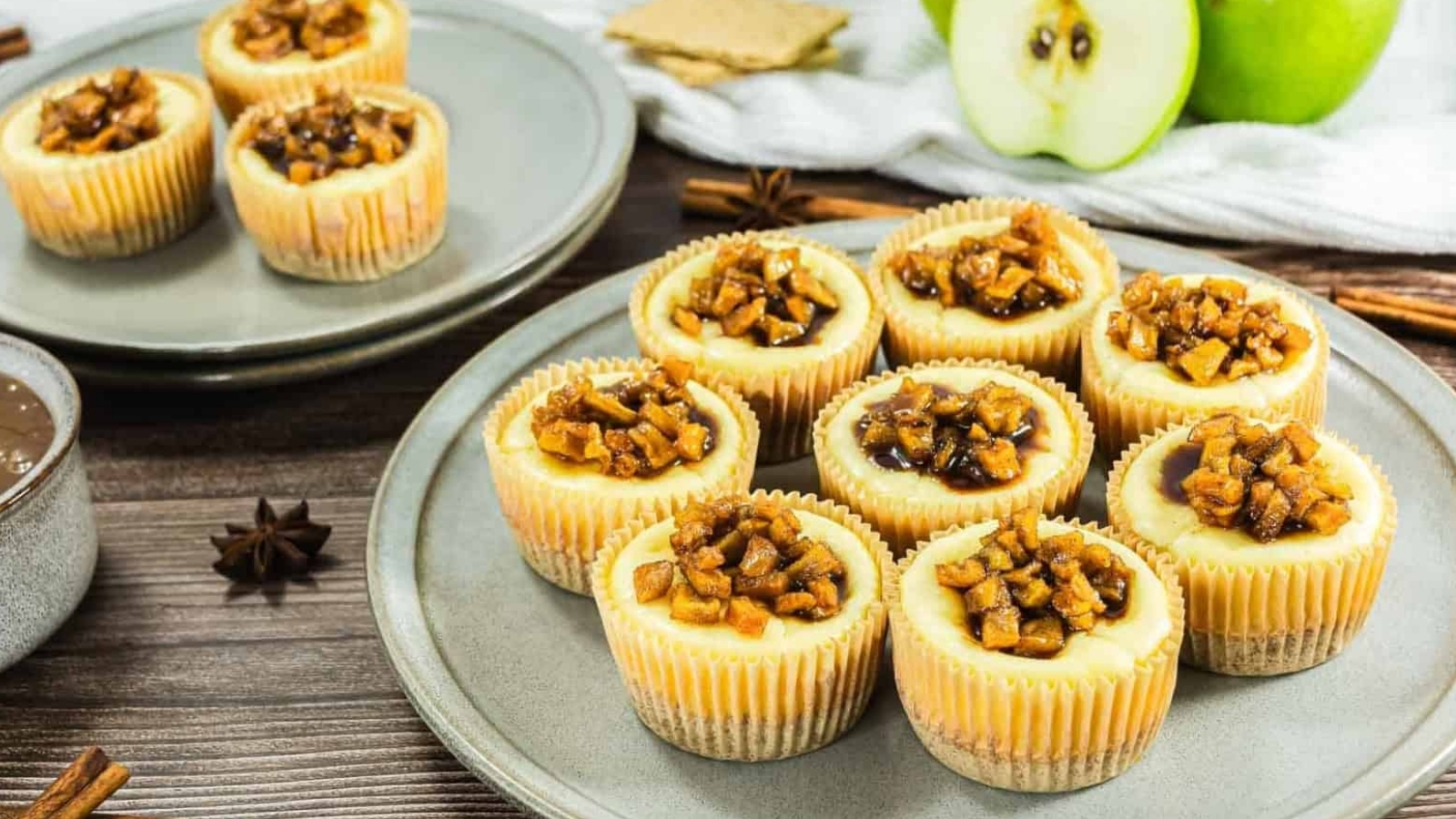 Mini cheesecakes topped with caramelized apples on plates, with a green apple and graham crackers in the background.