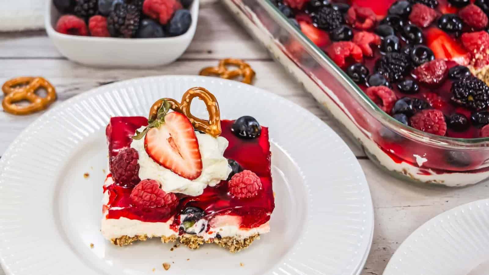 A slice of berry-topped dessert with a pretzel crust, whipped cream, and a strawberry half sits on a white plate. A glass dish with more dessert and a bowl of mixed berries are in the background.