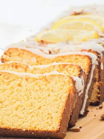 A loaf of lemon pound cake with icing, sliced on a wooden board, with lemons and almonds beside it.