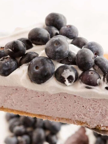 A slice of purple dessert with a crust and white topping covered in blueberries is held on a spatula above more blueberries.