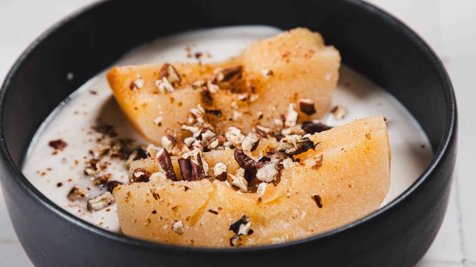 A bowl of yogurt topped with two peach slices, chopped nuts, and a sprinkle of cinnamon. The food is served in a dark bowl, placed on a light-colored surface.
