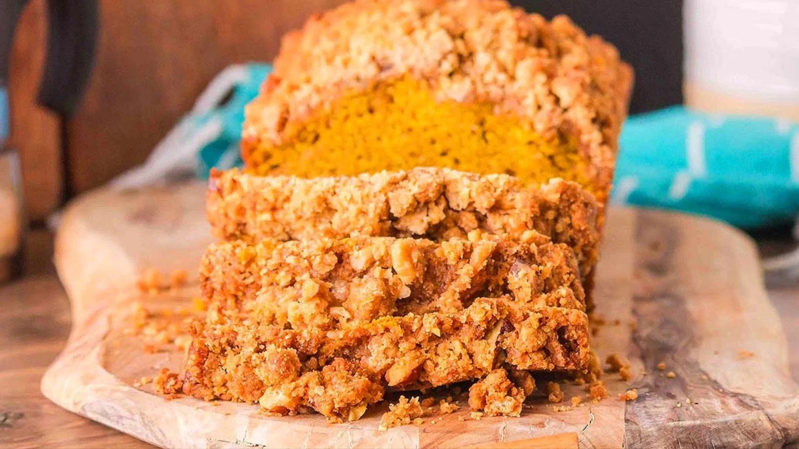 Loaf of pumpkin bread topped with a crunchy crumble topping, partially sliced on a wooden cutting board with visible pumpkin-colored interior.