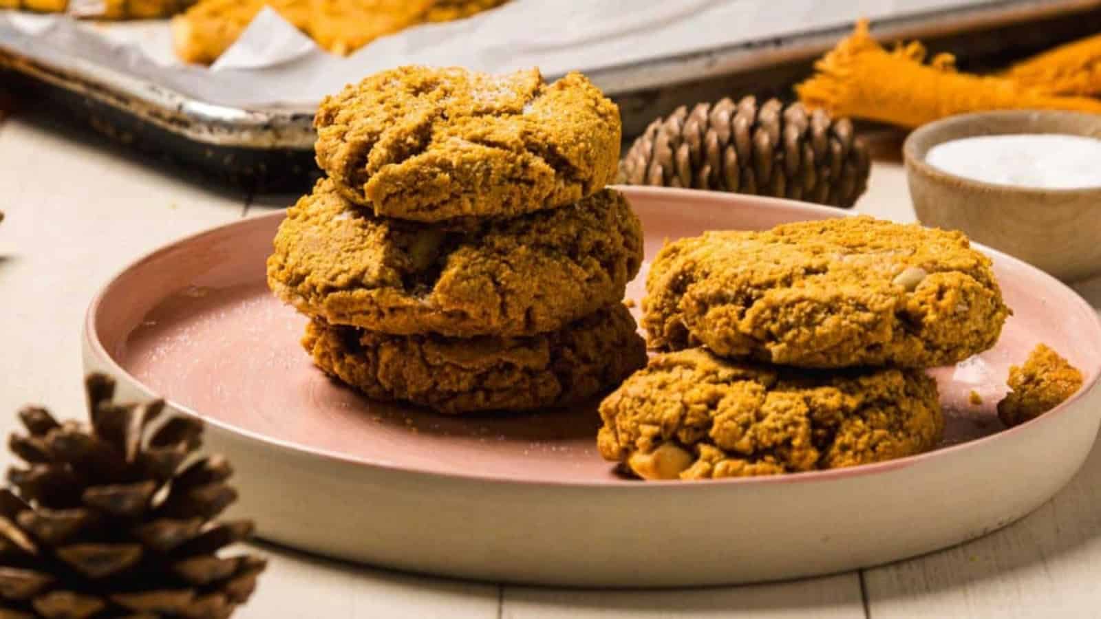 Four pumpkin cookies stacked on a pink plate, surrounded by pinecones and a baking tray in the background.