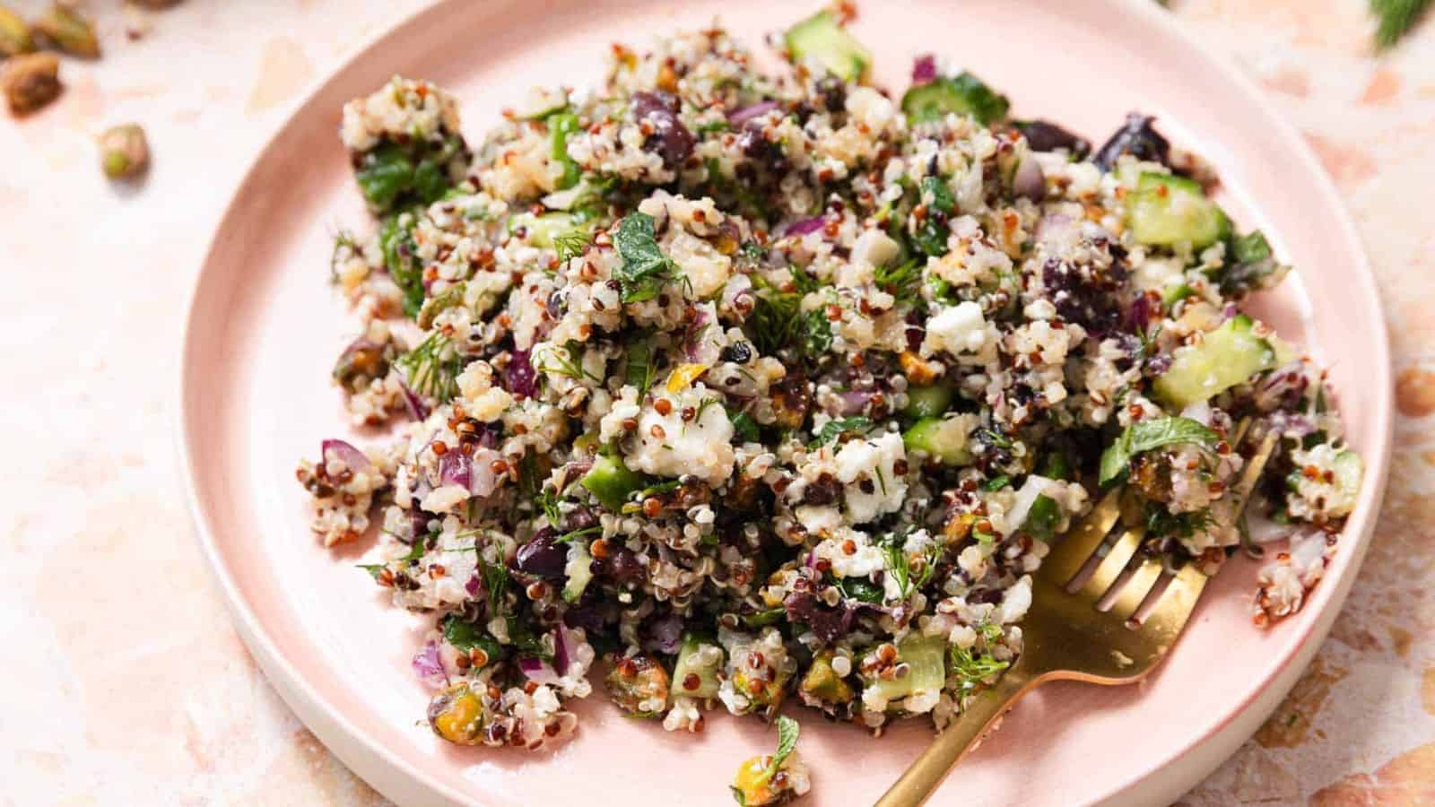 A white plate with a serving of quinoa salad mixed with diced cucumbers, chopped herbs, red onions, nuts, and feta cheese. A gold fork is resting on the plate. The background is a light, textured surface.