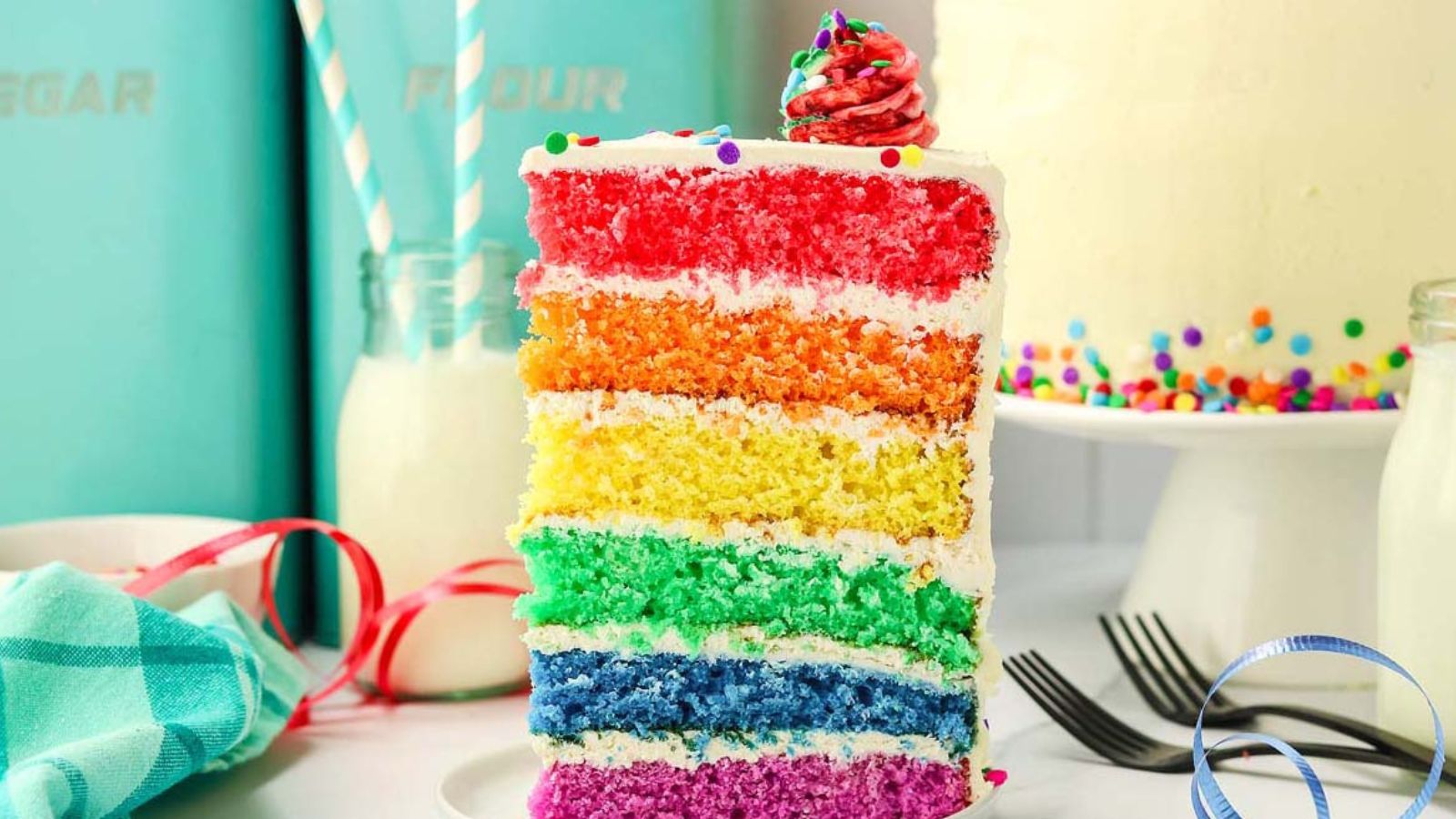 Slice of rainbow layer cake with white frosting and sprinkles on top, placed on a white plate with forks, a bowl of sprinkles, and two glass bottles with striped straws in the background.