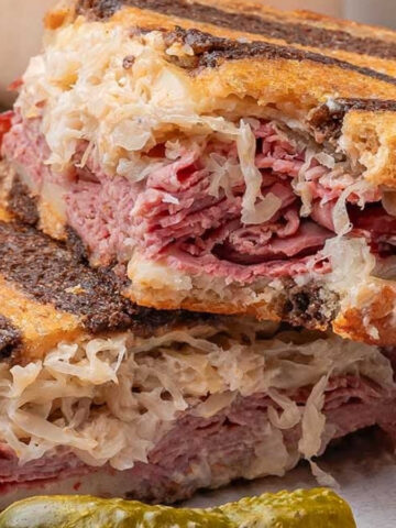 Two stacked grilled sandwiches with sliced meat and sauerkraut on rye bread, served with a pickle and a small bowl of sauce.