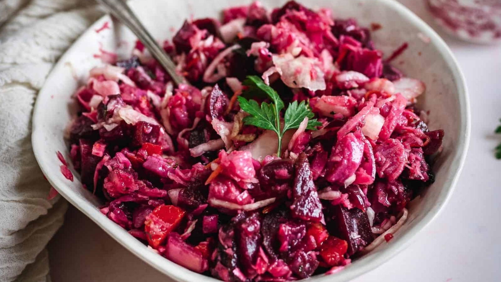 A bowl of chopped beet and cabbage salad with visible onion slices and a parsley leaf on top, with a spoon resting inside.
