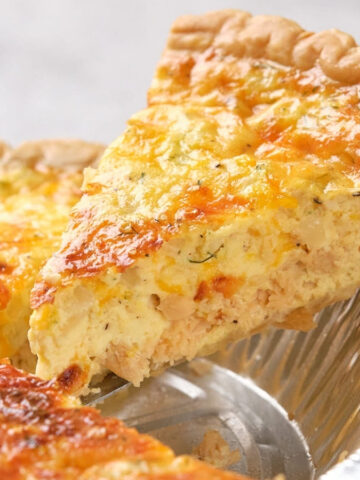 A hand lifting a slice of salmon quiche from a pie tin, revealing a golden, crispy top and creamy filling.