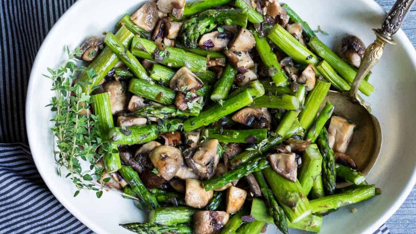 Chopped asparagus and sliced mushrooms are served in a white bowl with a spoon on a striped cloth.