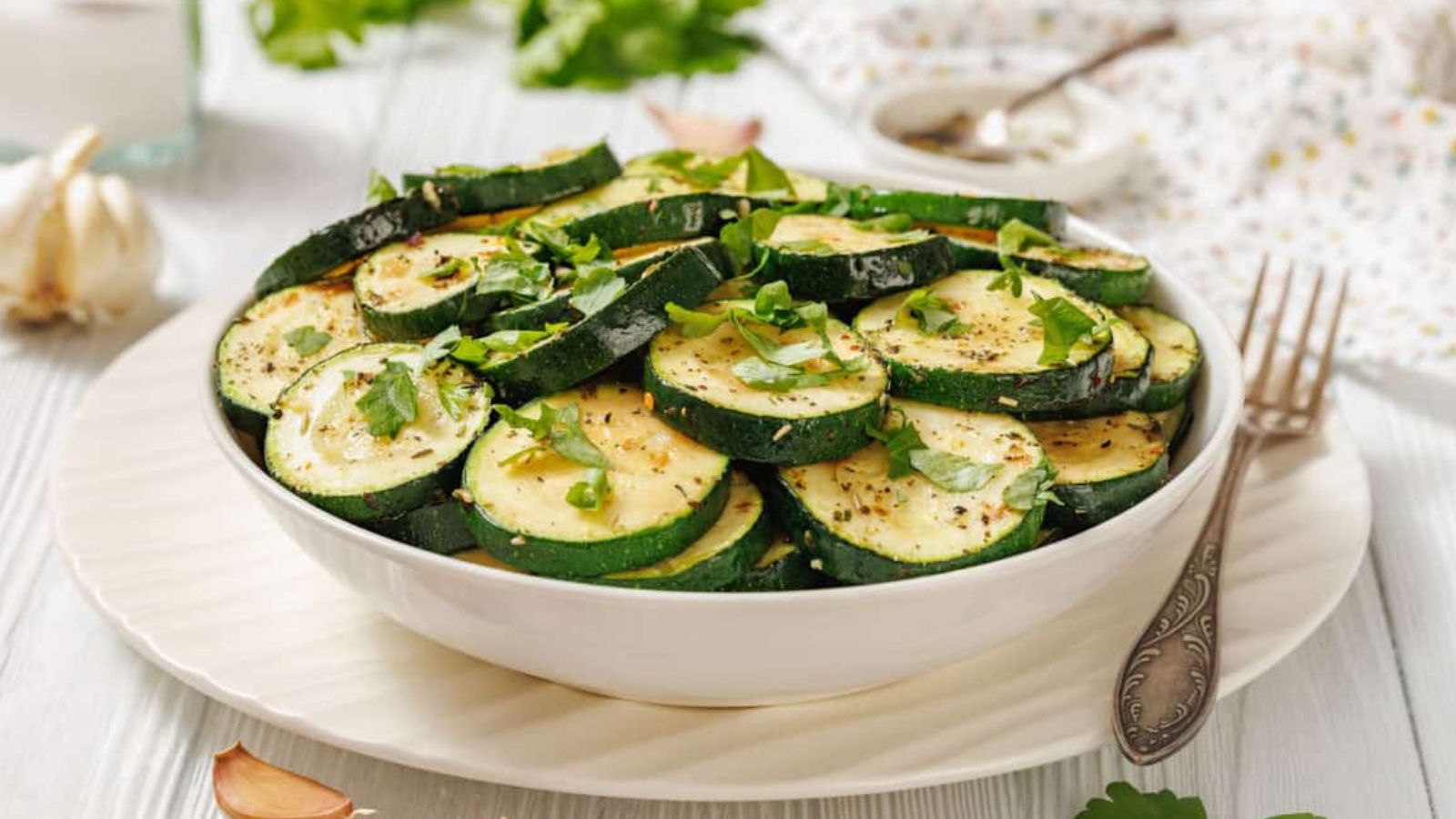 A white bowl filled with sliced Sautéed Zucchini with Garlic and Herbs, garnished with chopped herbs, is placed on a white plate with a fork nearby.