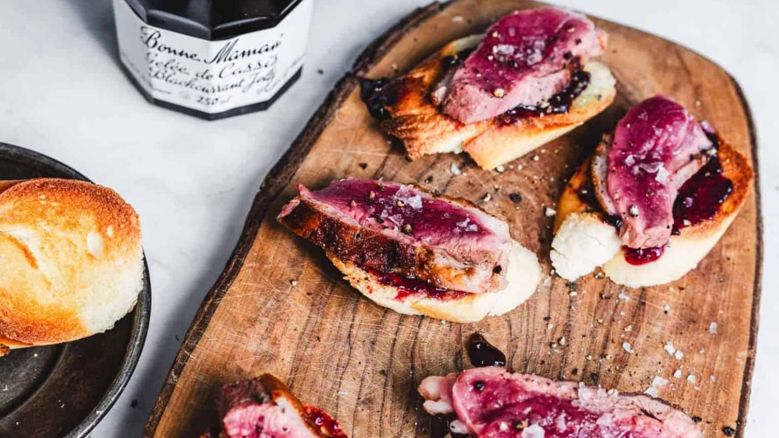Slices of beef on toasted bread with a dark berry sauce are arranged on a wooden serving board. A jar of Bonne Maman blackcurrant jelly is nearby. Another piece of plain toasted bread is on a plate to the side.