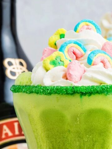 A tall glass of milkshake topped with whipped cream and colorful cereal marshmallows sits on a surface with additional marshmallows scattered around it.