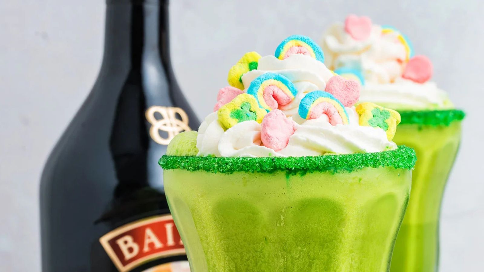 A tall glass of milkshake topped with whipped cream and colorful cereal marshmallows sits on a surface with additional marshmallows scattered around it.