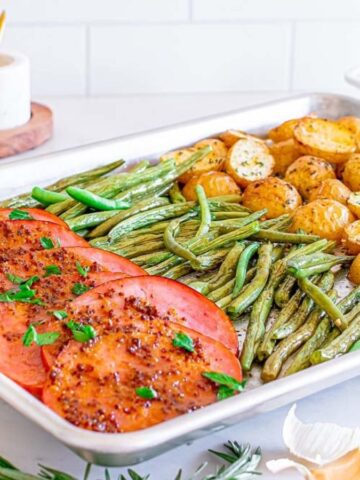 A sheet pan with sliced ham, green beans, and halved potatoes arranged in sections.