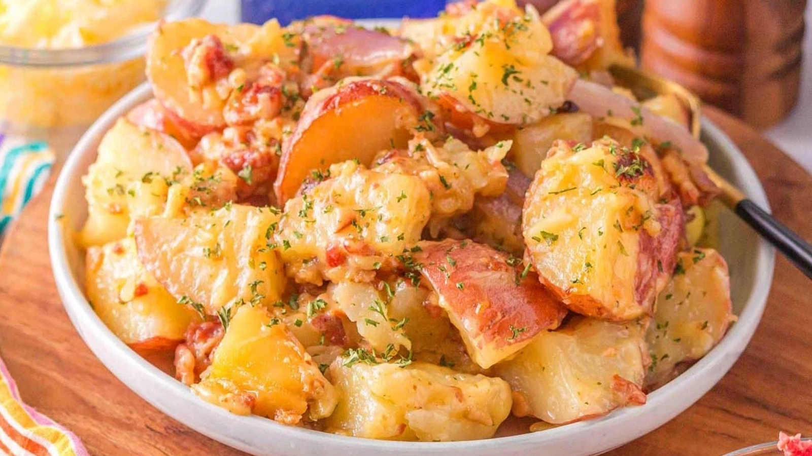 A bowl filled with chopped red-skinned potatoes mixed with melted cheese and herbs, with a spoon on the side.