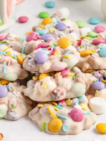 A pile of white-coated snack clusters topped with pastel candy-coated chocolates and sprinkles sits on a white surface with scattered candies, a glass bowl of candy pieces, and small marshmallows nearby.