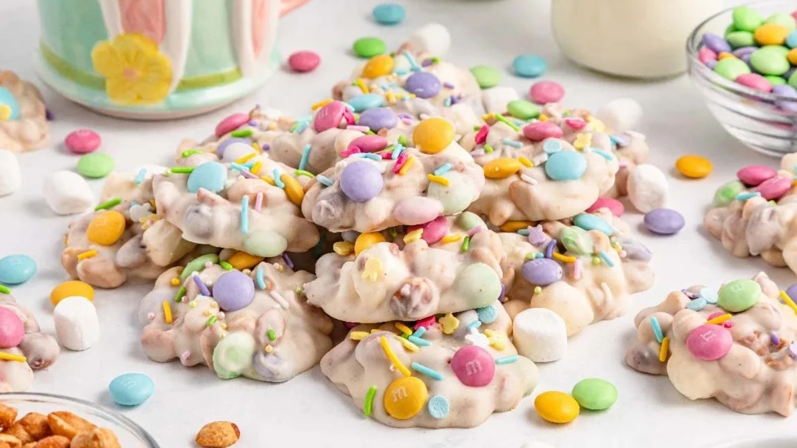 A pile of white-coated snack clusters topped with pastel candy-coated chocolates and sprinkles sits on a white surface with scattered candies, a glass bowl of candy pieces, and small marshmallows nearby.
