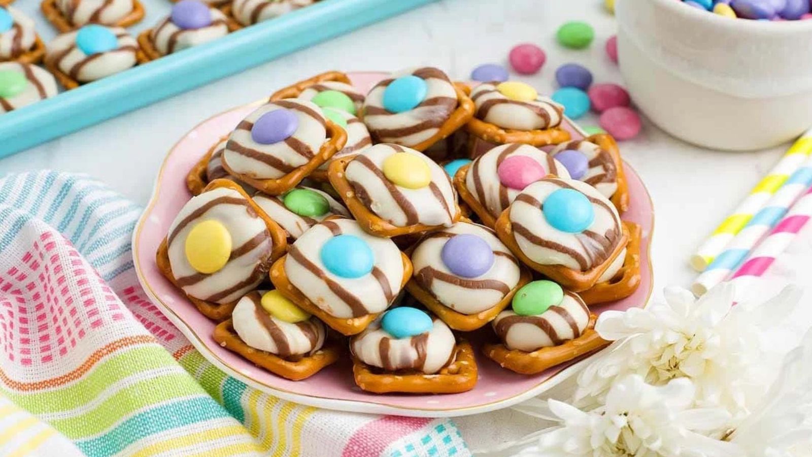 Pretzel squares topped with melted chocolate candies and pastel candy-coated chocolates arranged like flowers.