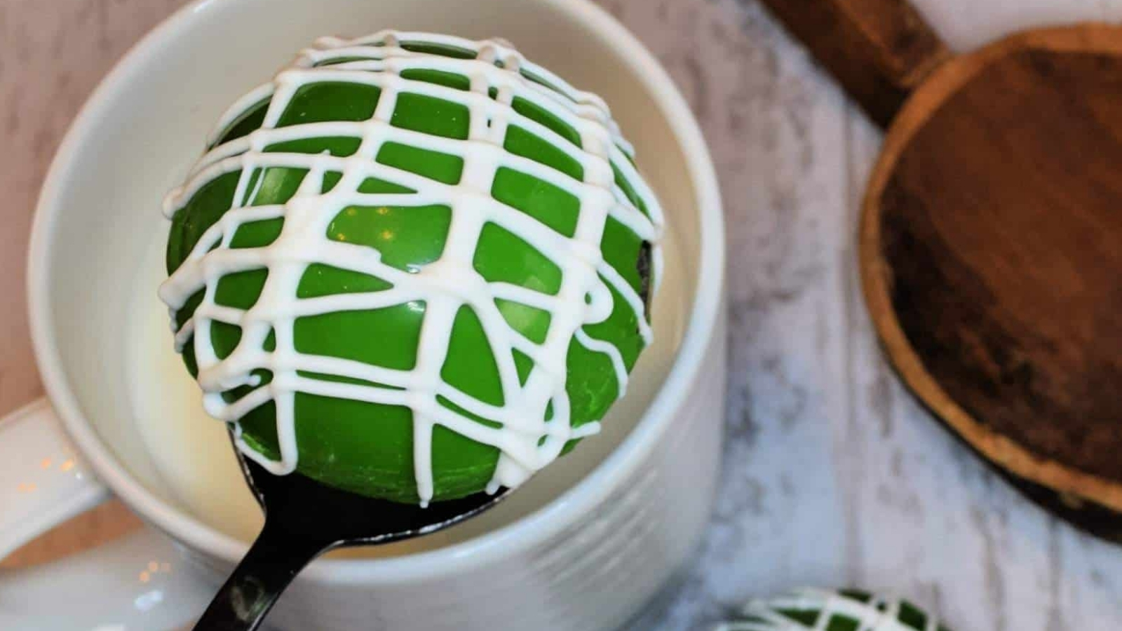 St. Patrick's Day Hot Chocolate Cocoa Bomb with white icing drizzle, resting on a spoon over a white cup.