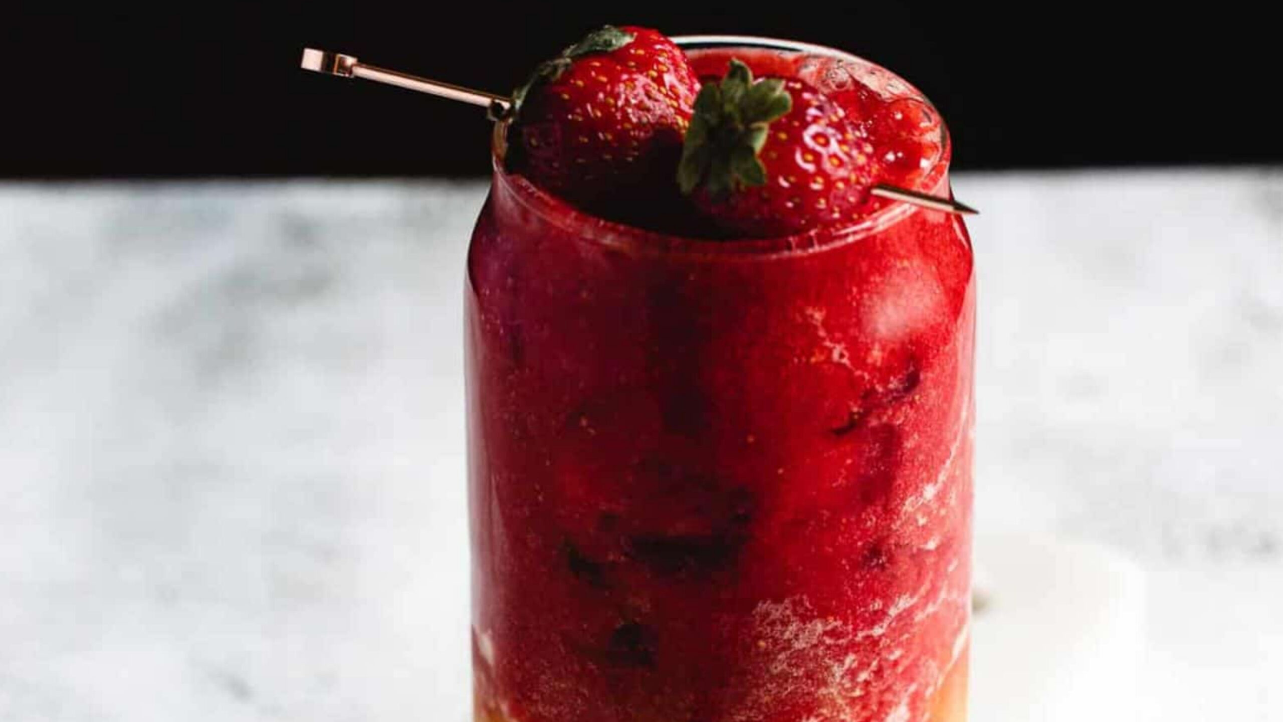 A glass filled with a red slushy drink, topped with whole strawberries on a skewer. The surface below is light and slightly textured, contrasting with the dark background.