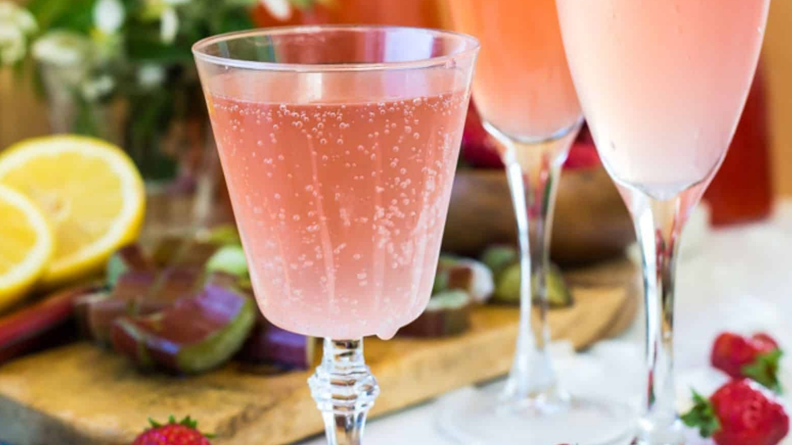 A glass filled with a bubbly pink drink is in focus. In the background, there are two more glasses with similar drinks. Cut lemons, strawberries, and flowers are also visible on a wooden board.
