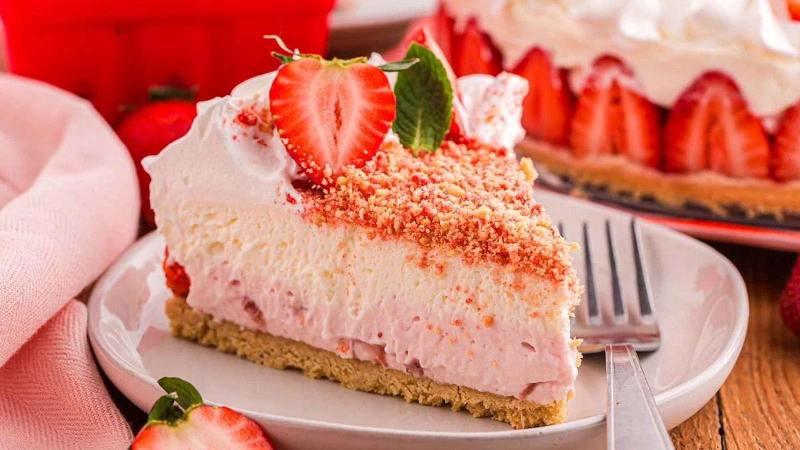 A slice of strawberry cream dessert with a crumb crust sits on a plate, topped with a halved strawberry and crumbs, with a larger matching dessert in the background.