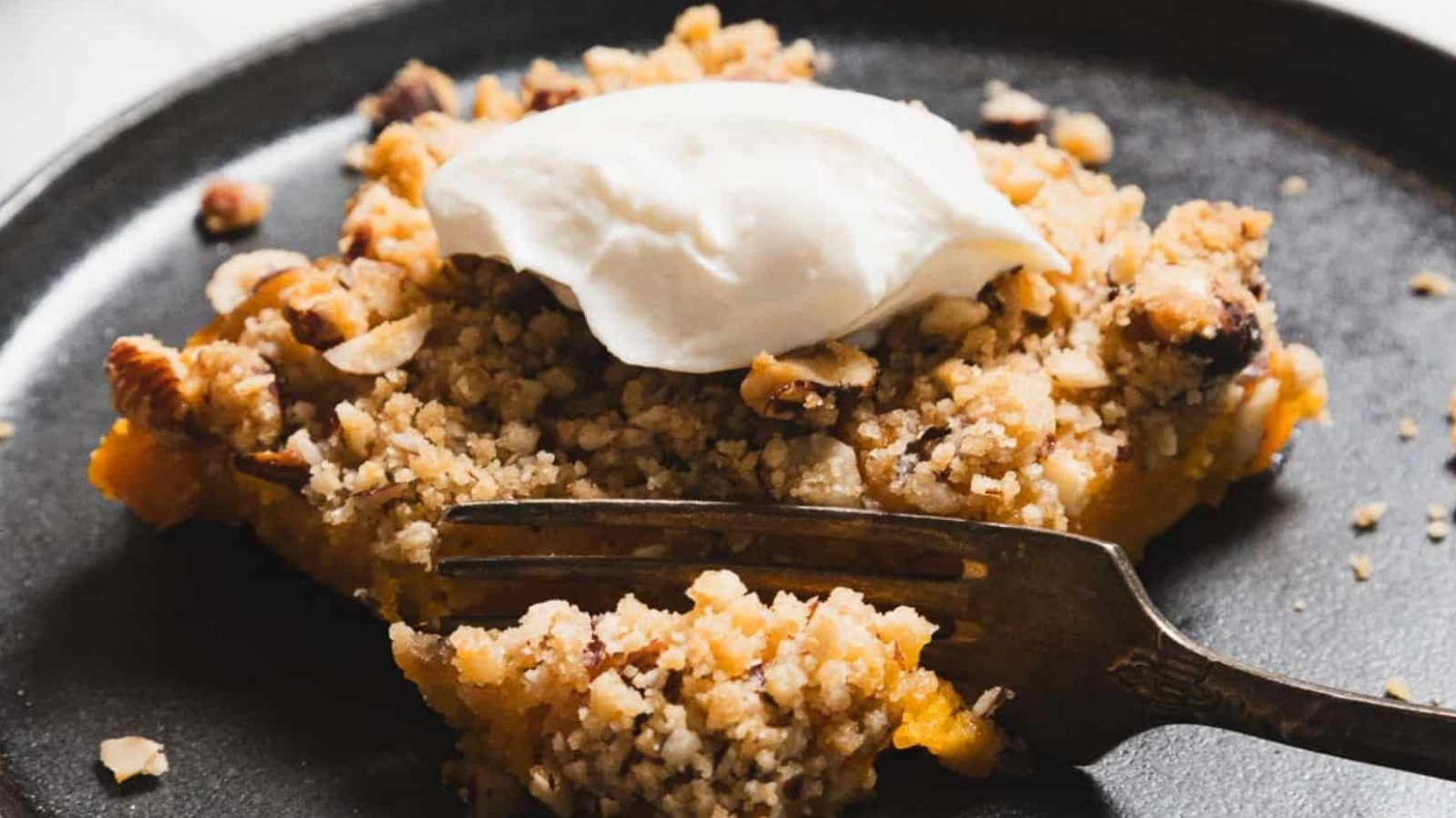 A slice of crumbly dessert topped with a dollop of white cream is presented on a dark plate. The dessert is garnished with chopped nuts, and a fork rests in front of it, partially obscuring the dessert.