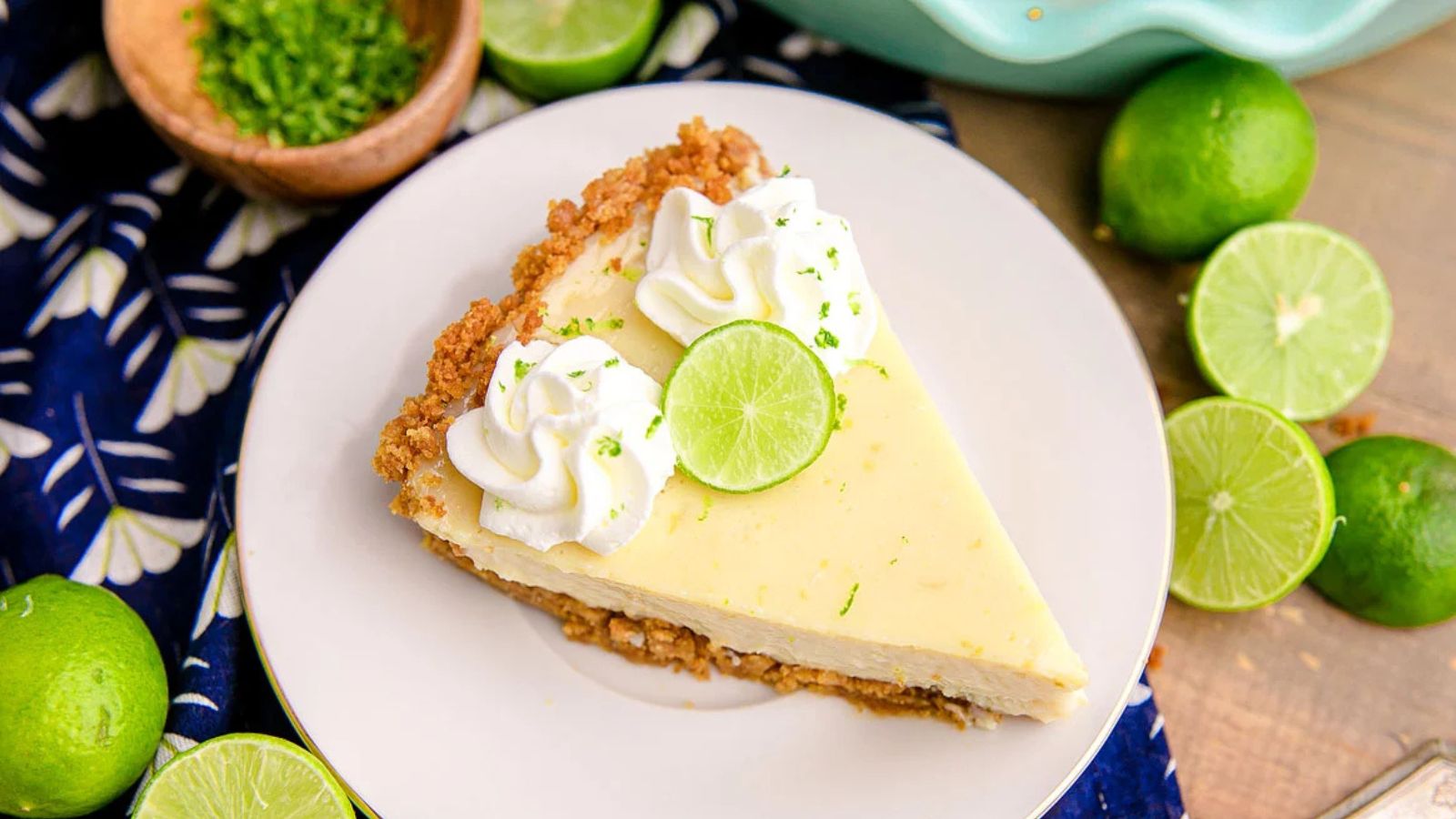 Slice of key lime pie with whipped cream and a lime slice on top, served on a white plate surrounded by whole and sliced limes.