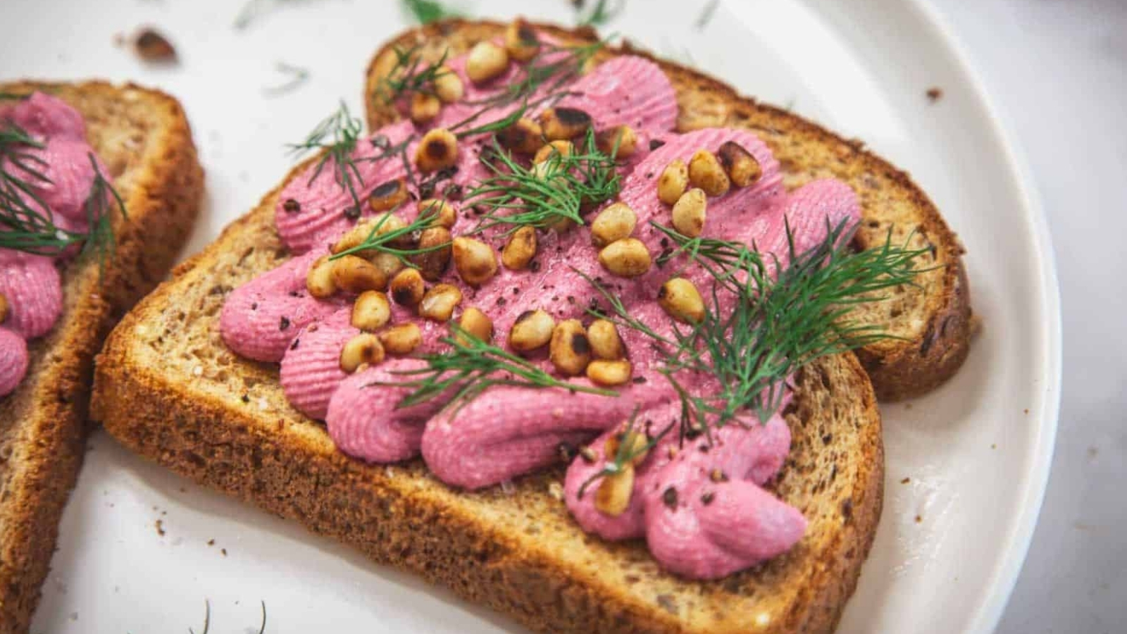 Two slices of whole grain toast topped with piped pink spread, toasted pine nuts, and fresh dill, served on a white plate.