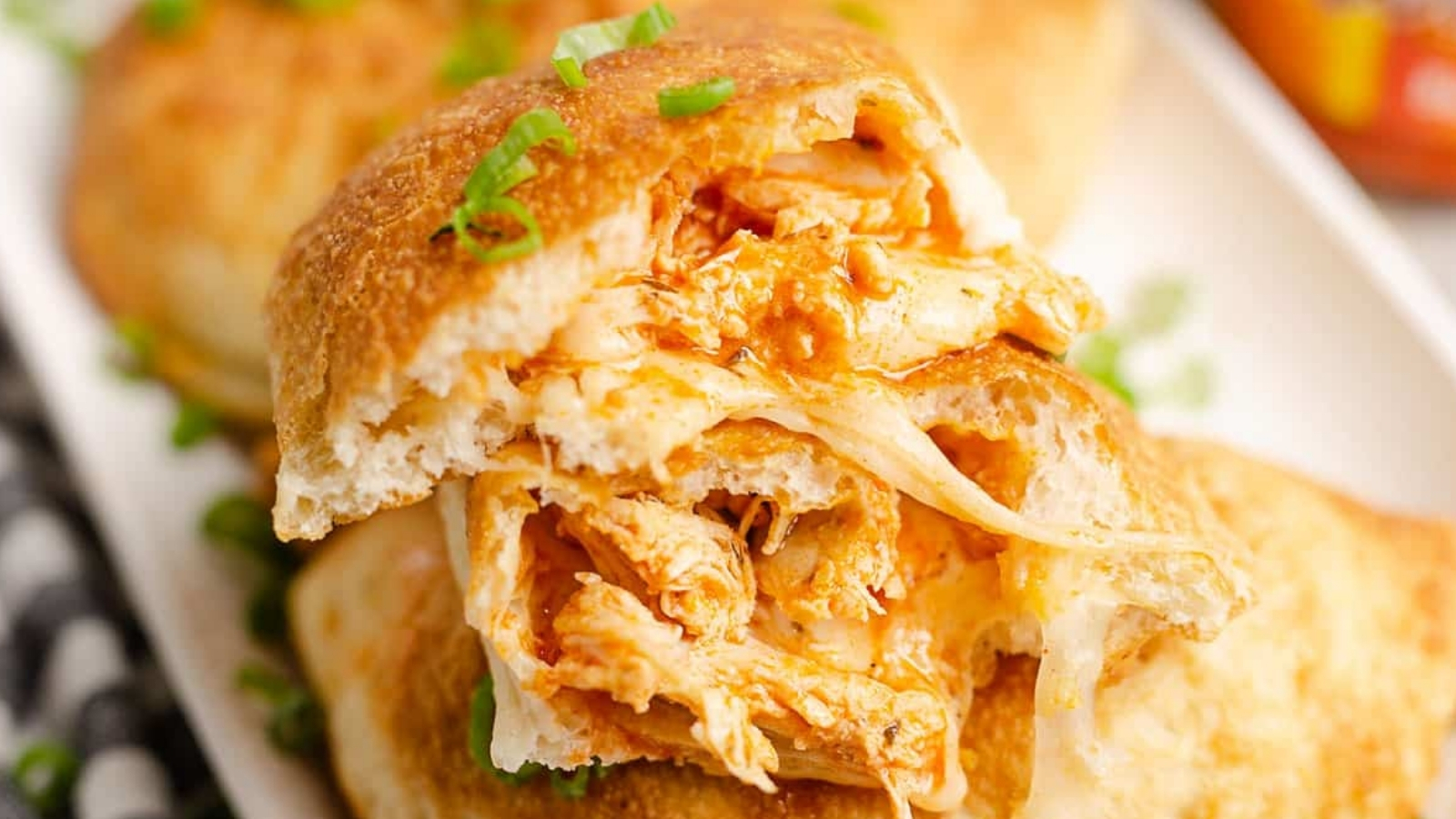 A sandwich with shredded chicken and melted cheese inside a toasted bun, topped with chopped green onions on a white plate.