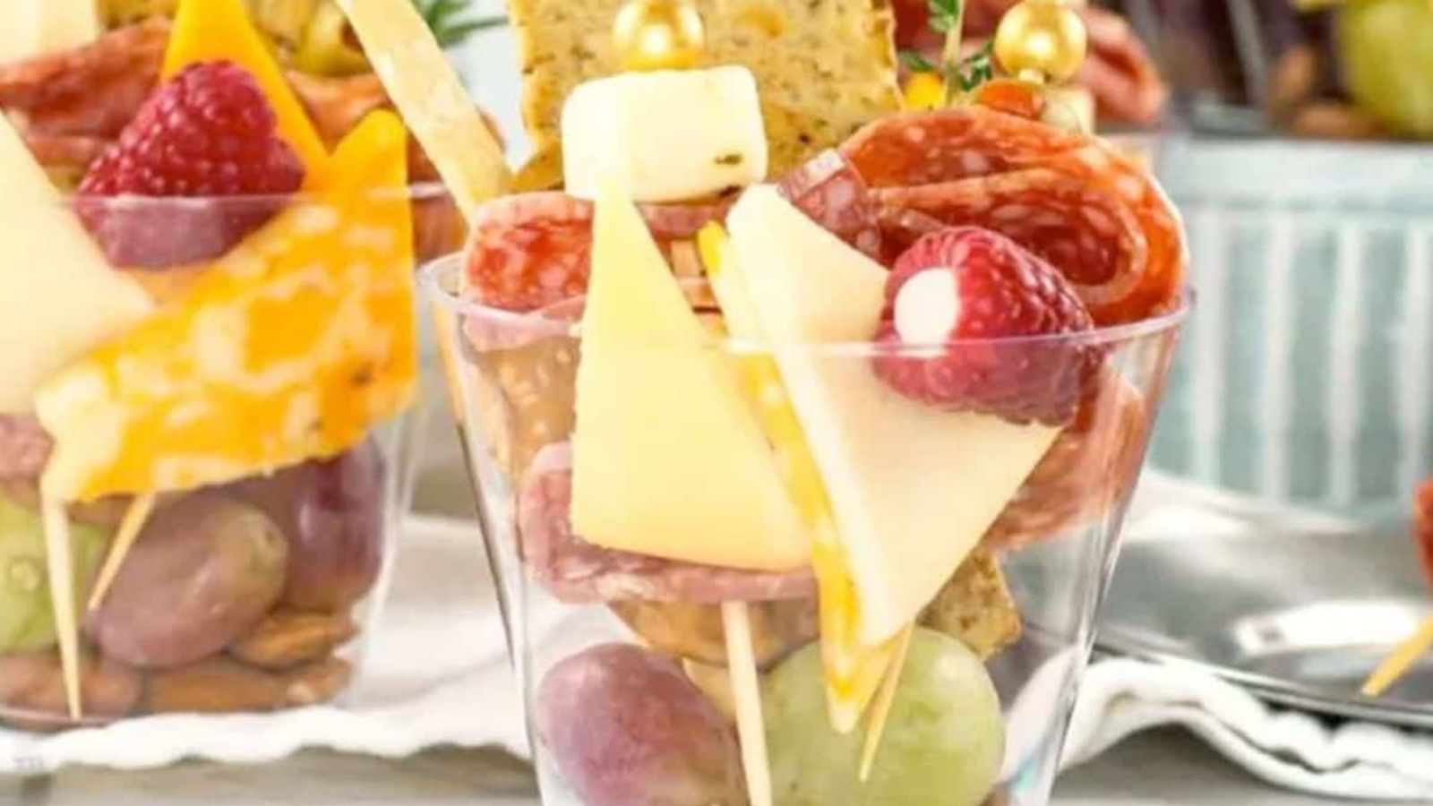 A clear plastic cup filled with grapes, almonds, cheese cubes, salami slices, a raspberry, and crackers, arranged on skewers for a single-serve charcuterie snack.