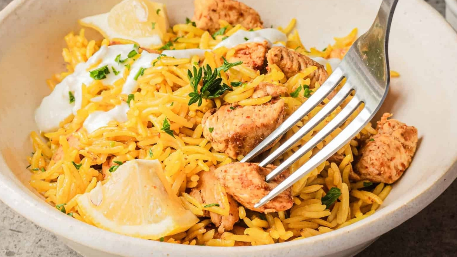 A fork in a bowl of yellow rice with grilled chicken, lemon wedges, herbs, and a dollop of white sauce.