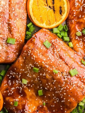 Glazed salmon fillets on a tray topped with sesame seeds and chopped green onions, surrounded by sliced oranges.