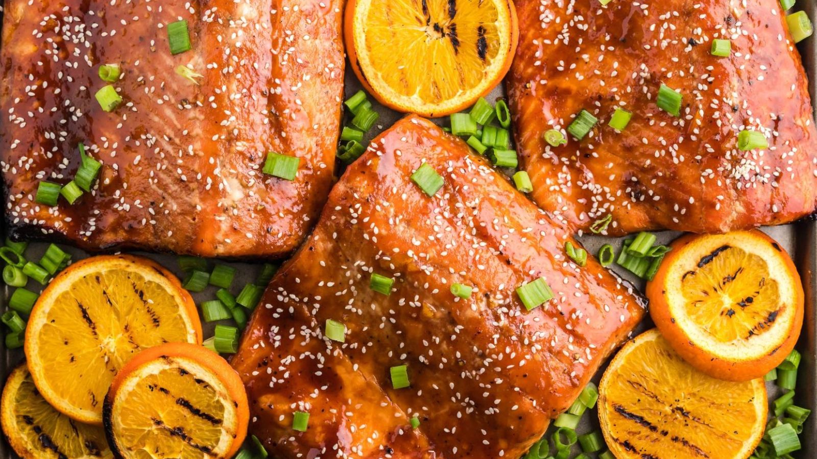 Glazed salmon fillets on a tray topped with sesame seeds and chopped green onions, surrounded by sliced oranges.