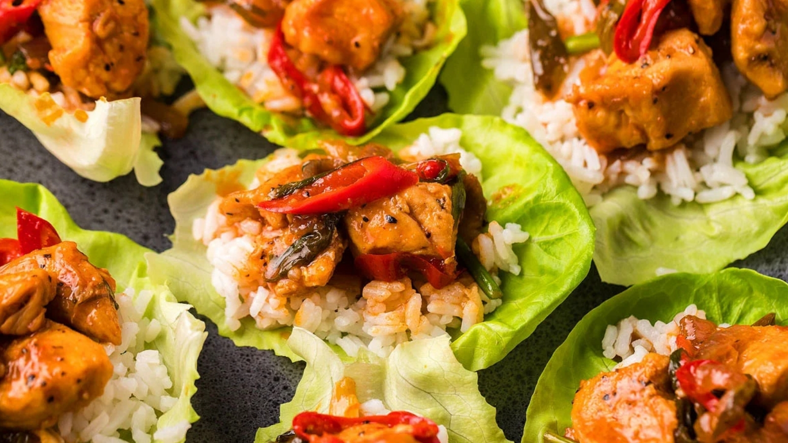 Lettuce leaves filled with white rice and pieces of chicken topped with sliced red peppers, arranged on a dark surface.