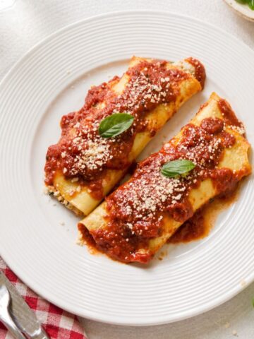 Homemade manicotti made with crepes instead of pasta shells topped with marinara sauce and grated Parmesan cheese on a white plate.