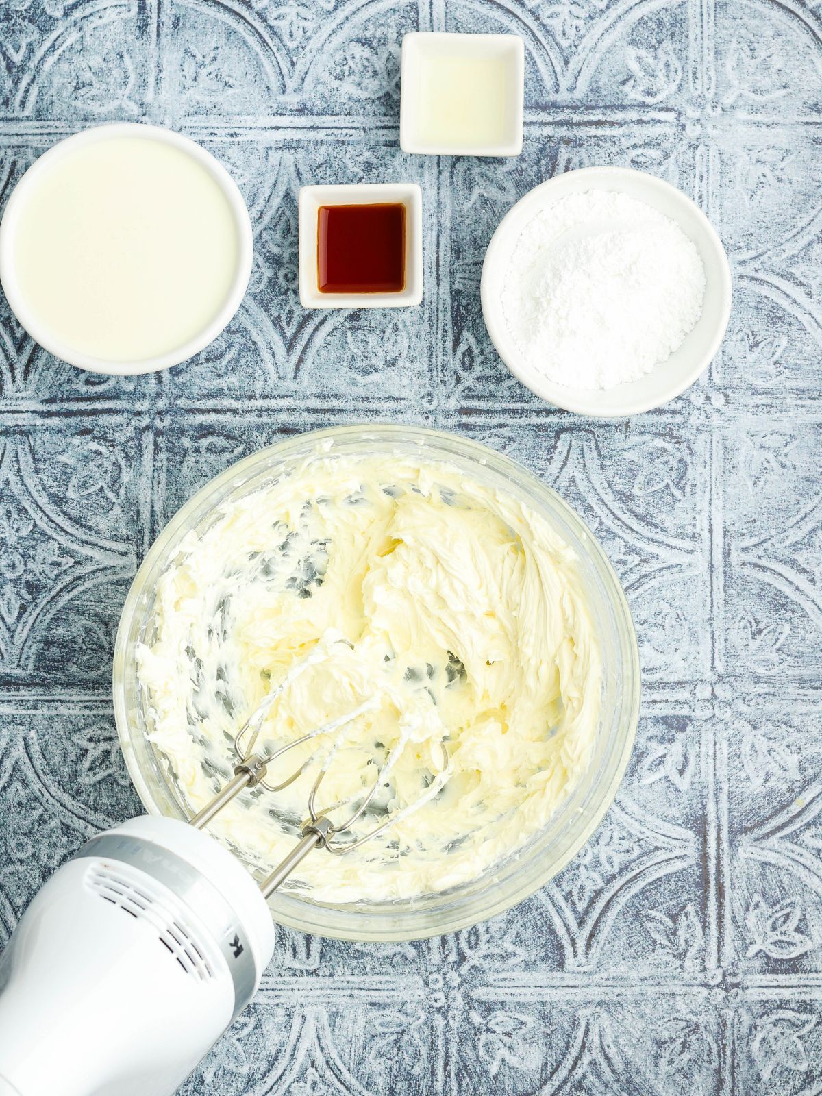 Overhead photo of sweetened cream cheese filling for dessert crepes in a glass mixing bowl with powdered sugar, vanilla, lemon extract, and cream on a patterned countertop.