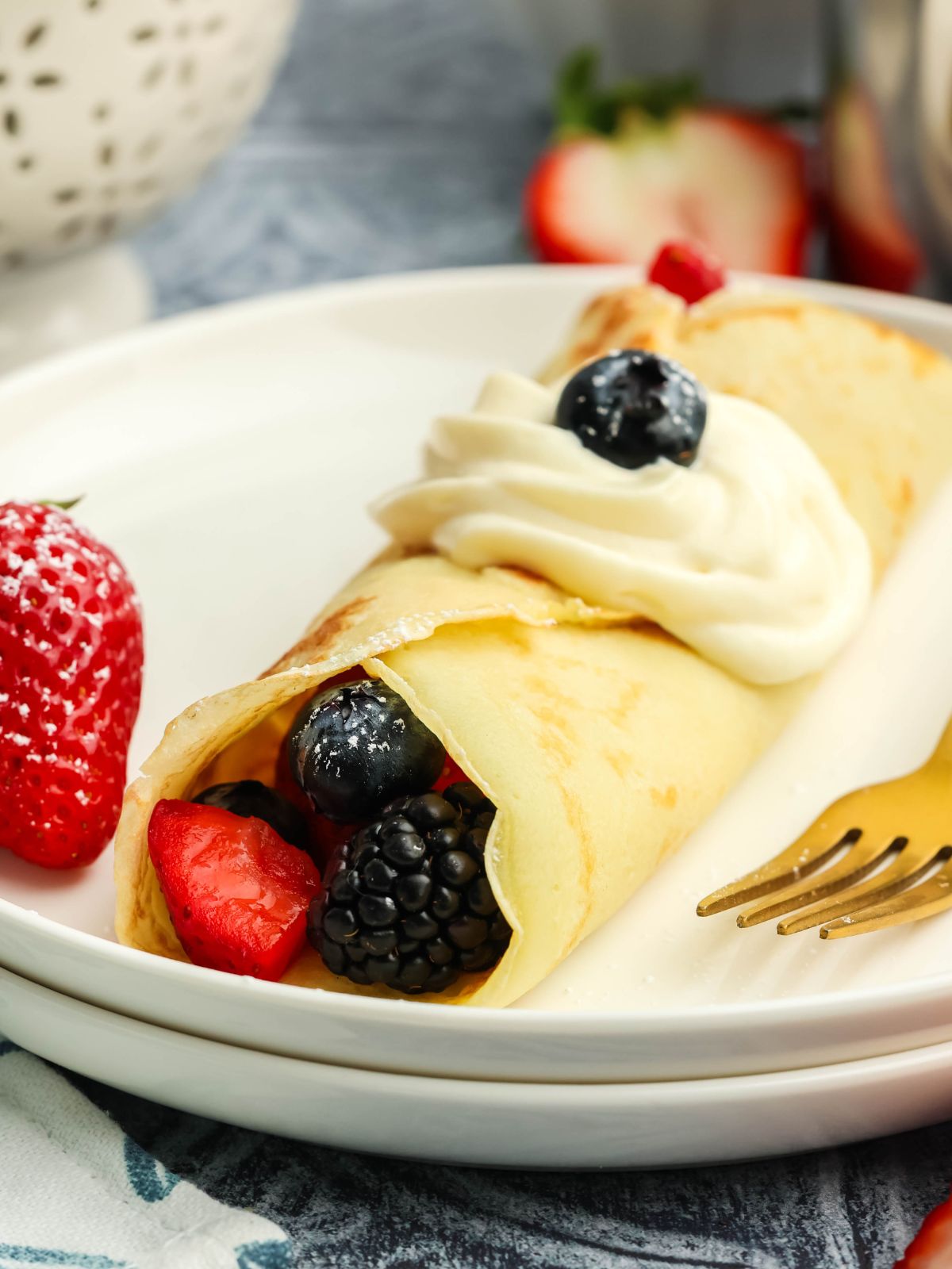Close-up photo of a dessert crepe filled with strawberries, blueberries, and blackberries topped with cream cheese filling on a white plate with a gold fork.