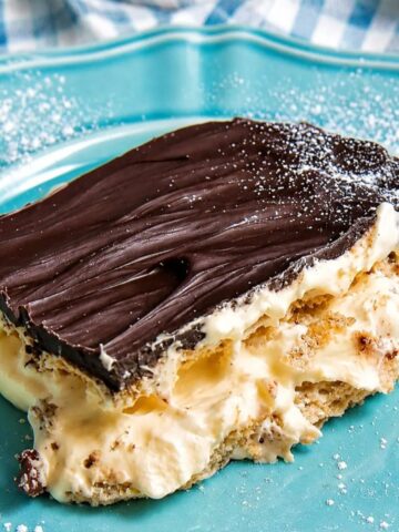 Slice of no bake chocolate eclair pie with layers of vanilla pudding, graham crackers, and chocolate ganache on a blue plate.