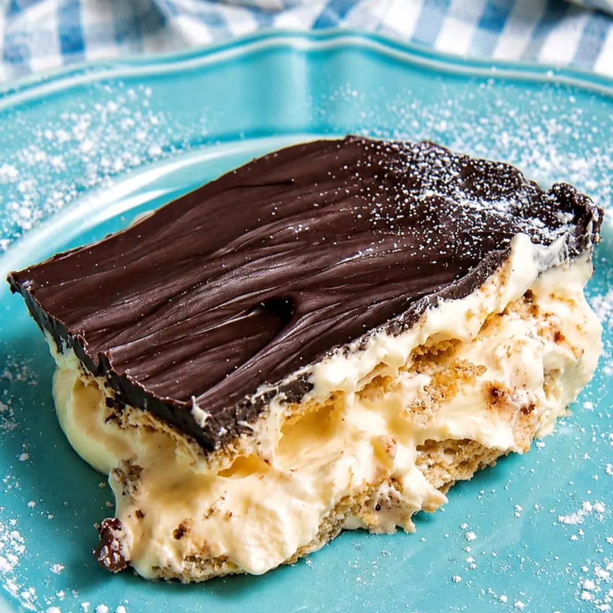 Slice of no bake chocolate eclair pie with layers of vanilla pudding, graham crackers, and chocolate ganache on a blue plate.