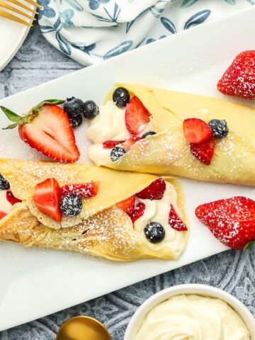 Dessert crepes filled with sweetened cream cheese, strawberries, and blueberries topped with powdered sugar on a white serving platter with fresh berries and gold serving spoons.