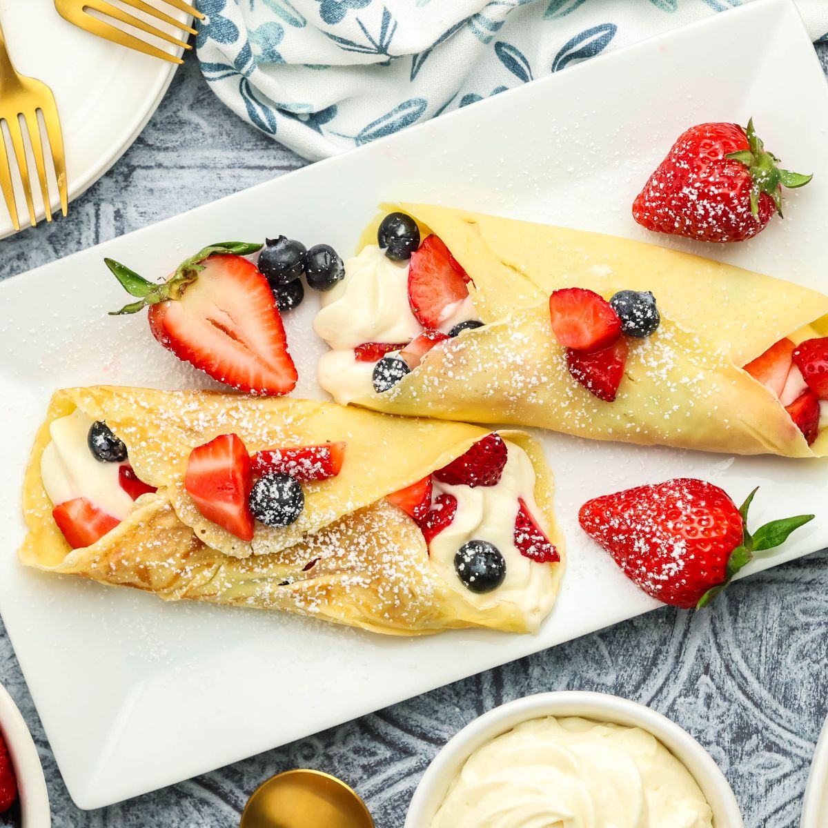 Dessert crepes filled with sweetened cream cheese, strawberries, and blueberries topped with powdered sugar on a white serving platter with fresh berries and gold serving spoons.