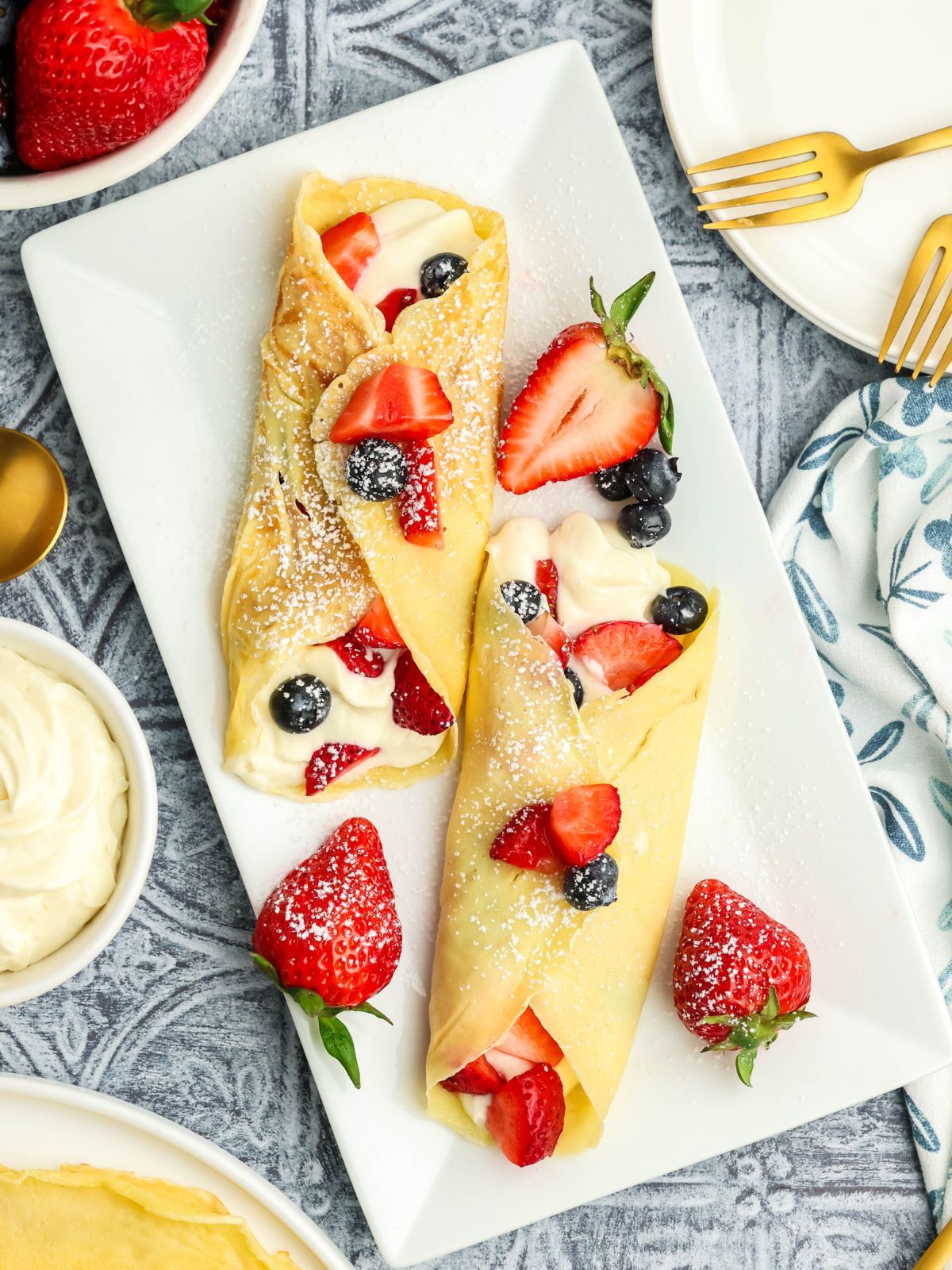 Dessert crepes filled with cream cheese filling, strawberries, and blueberries topped with powdered sugar on a white serving platter.