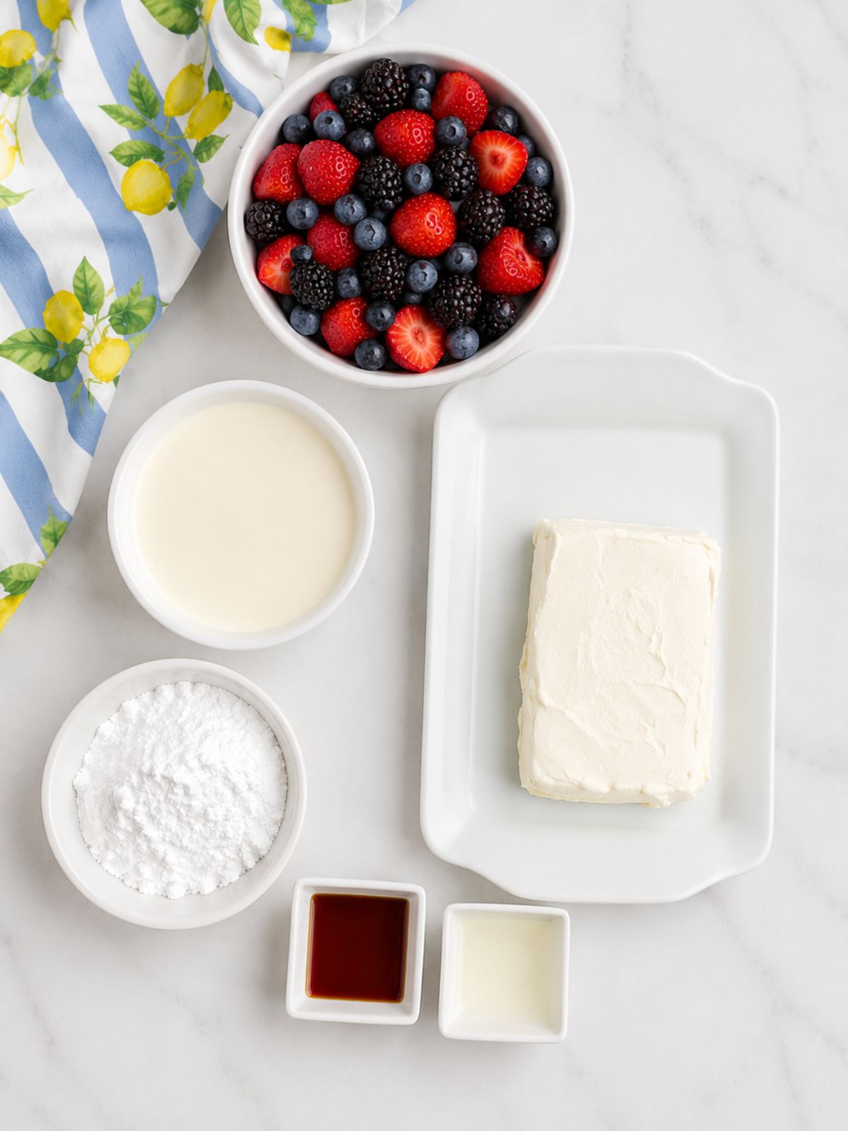 Overhead ingredient photo for dessert crepes featuring strawberries, blueberries, blackberries, cream cheese, powdered sugar, milk, vanilla, and lemon extract on a white marble countertop with a lemon-print kitchen towel.