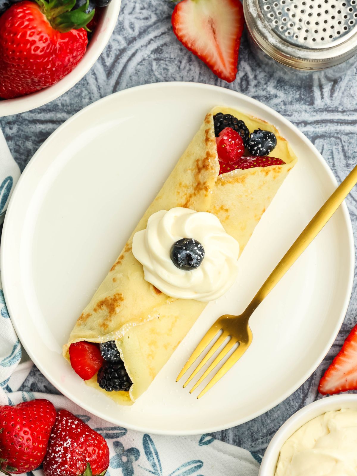 Dessert crepe filled with strawberries, blueberries, blackberries, and cream cheese filling topped with whipped cream on a white plate with a gold fork.