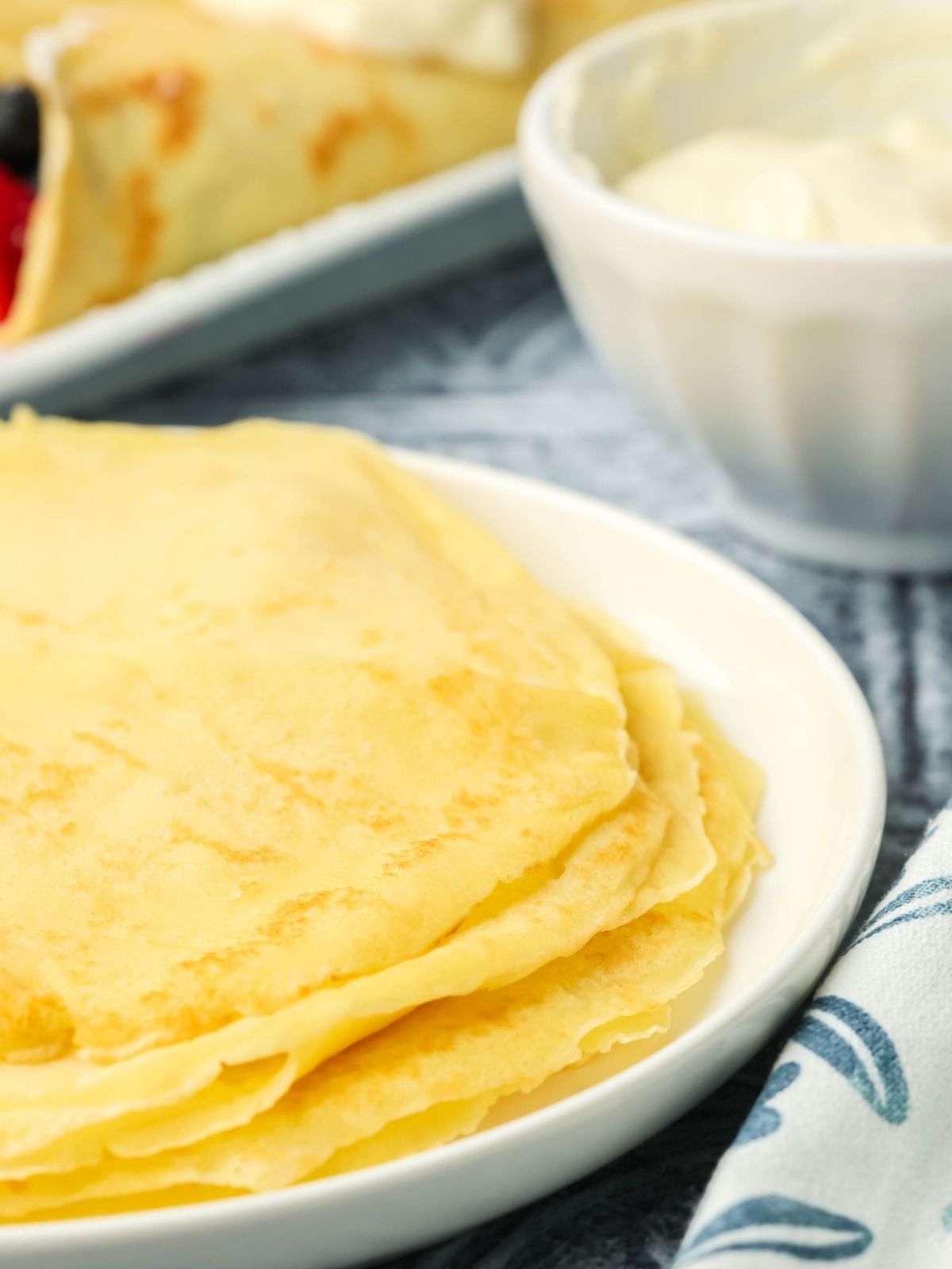 Stack of homemade crepes on a white plate with cream cheese filling and berry crepes blurred in the background on a kitchen countertop.
